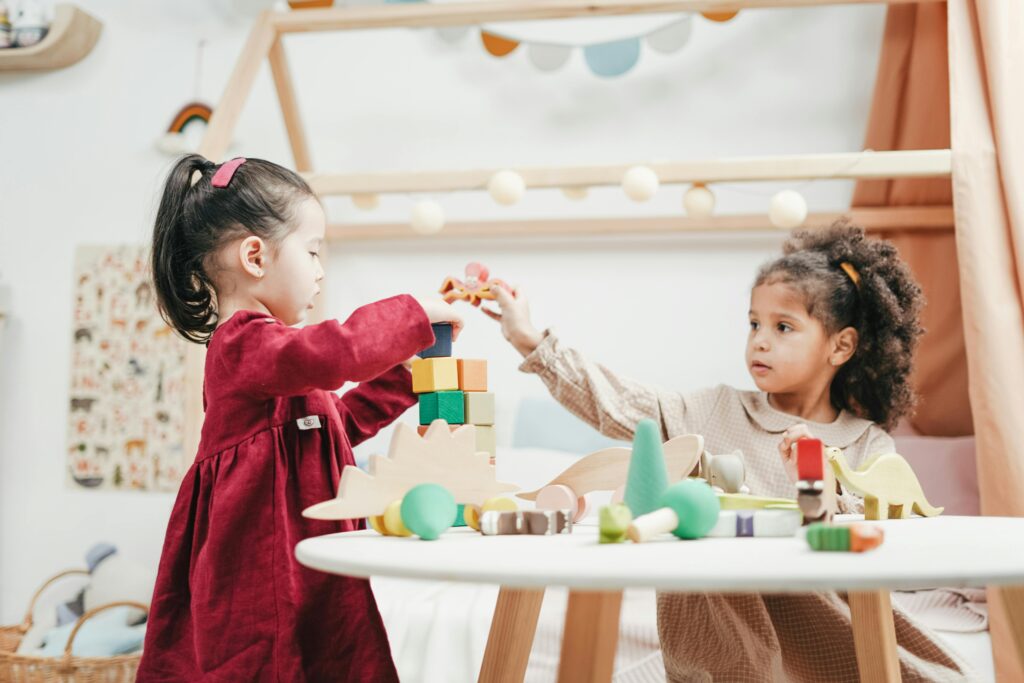Two young children playing with colorful building blocks at a round table in a bright, cozy room.