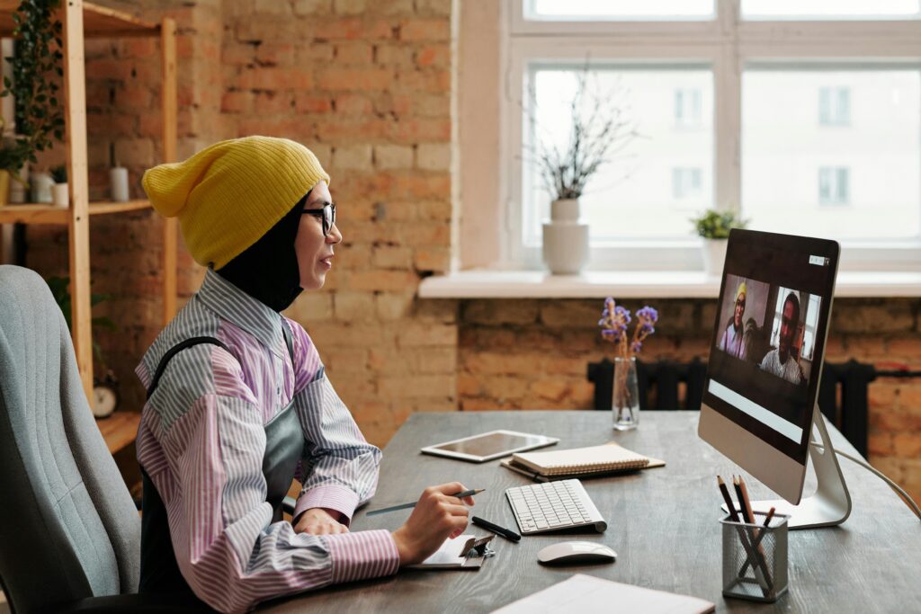 Person in a yellow beanie and glasses working on a computer at a desk with a notepad, near a window with plants.