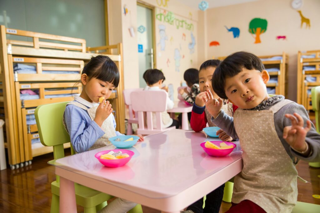 Children sitting at a table in a colorful classroom, eating from bowls and with decorative wall art in the background.