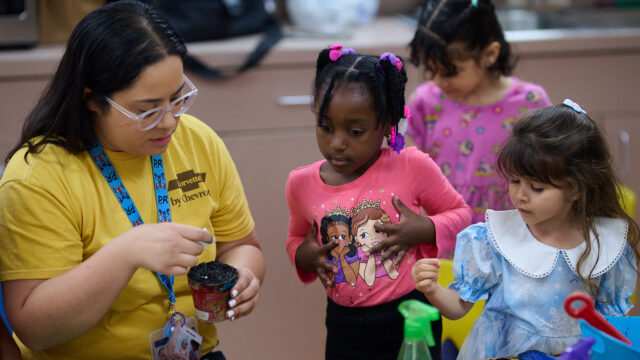 Teacher helping young children with a craft project at a classroom table, one child wearing a pink shirt, others nearby.