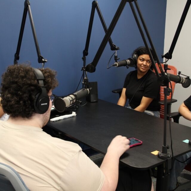 People recording in a podcast studio with microphones and headphones, sitting around a table against a blue wall.
