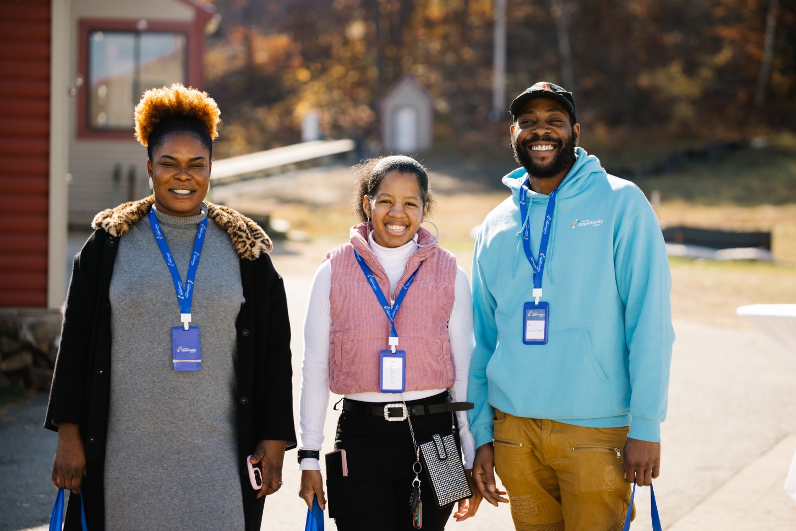 Three people standing outside, smiling, each wearing a badge, with autumn trees in the background.