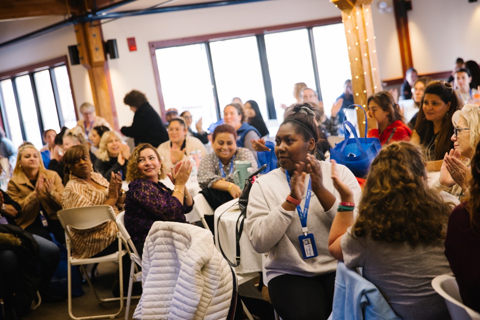 Diverse group of people sitting at tables in a bright room, clapping and engaging in a lively discussion.