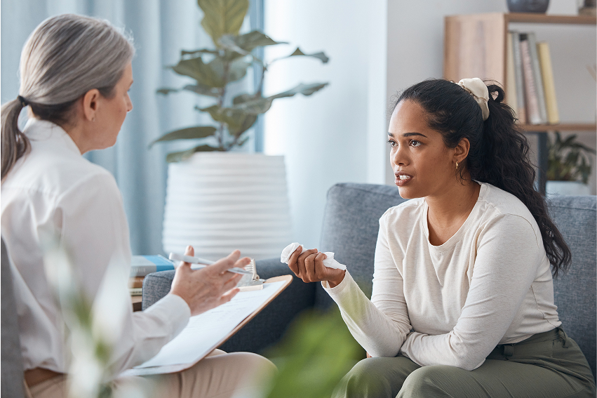 Two women having a serious conversation in a cozy room, one taking notes, the other holding a cup, plants and books nearby.