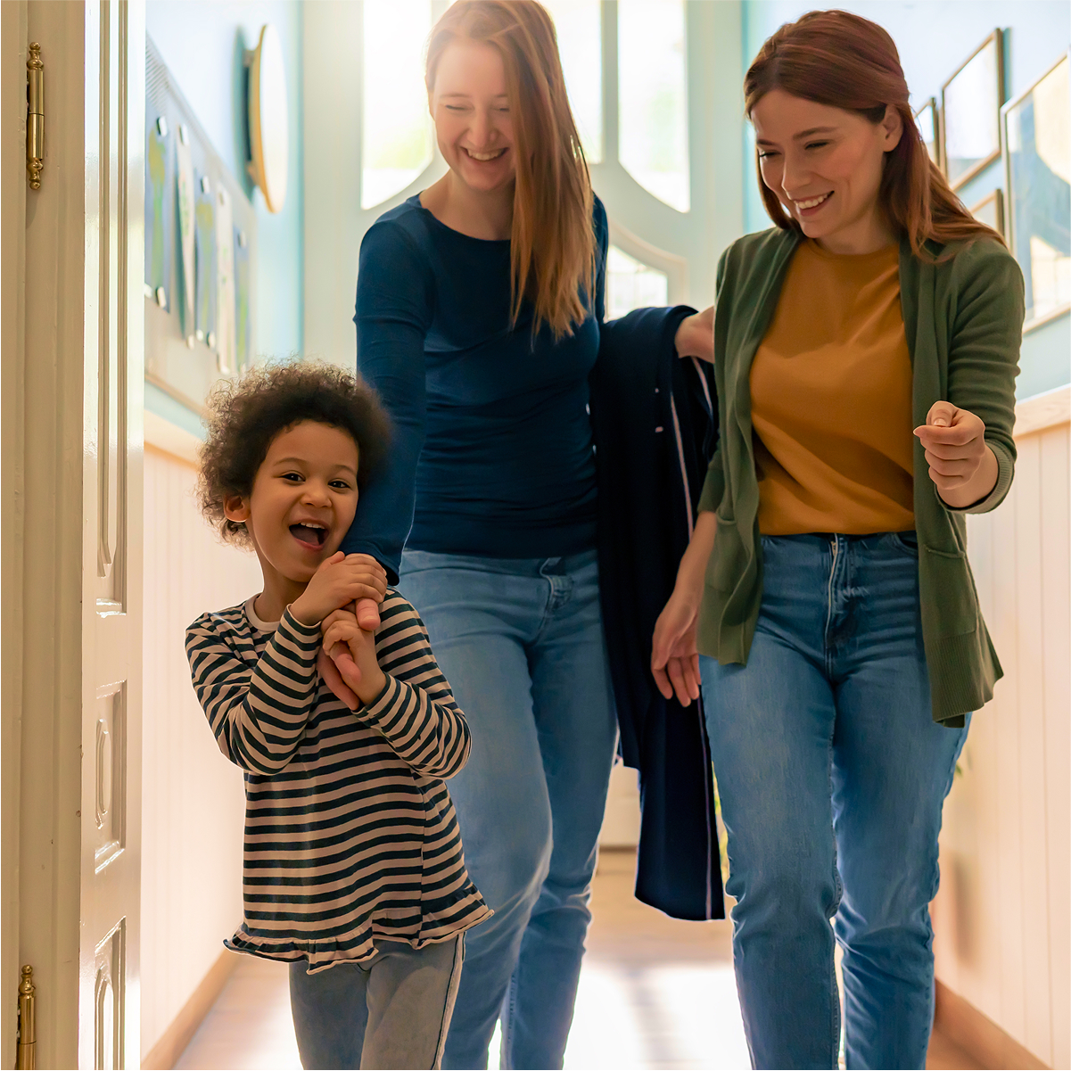 Child walking down a hallway with two smiling adults in casual clothing, brightly lit setting with framed pictures on the wall