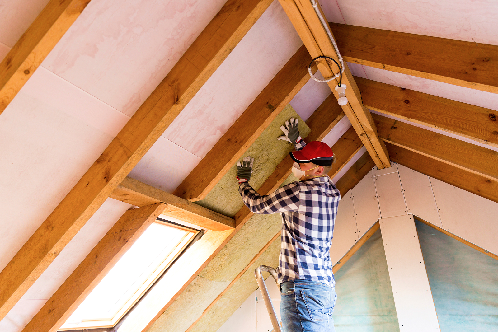 Person installing insulation in a wooden attic, wearing a plaid shirt and red cap, securing energy efficiency.