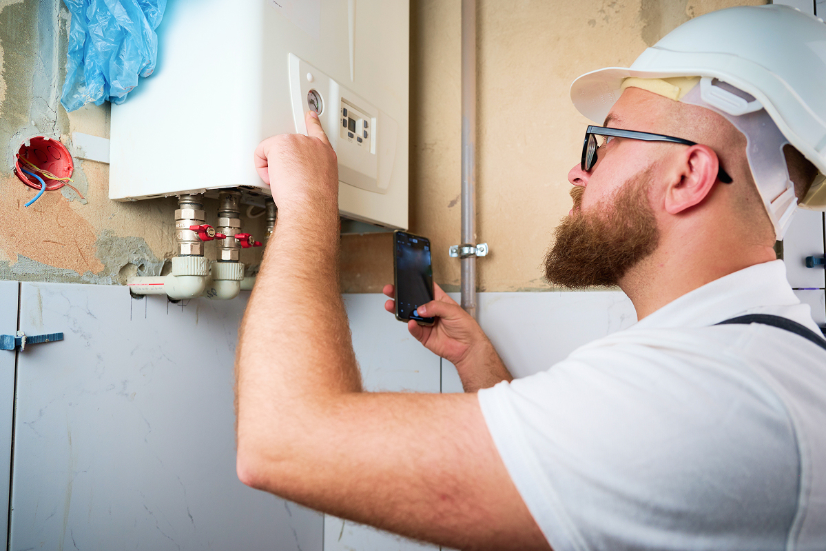 Plumber inspecting a water heater with a smartphone and tools in a utility room wearing a white hard hat.