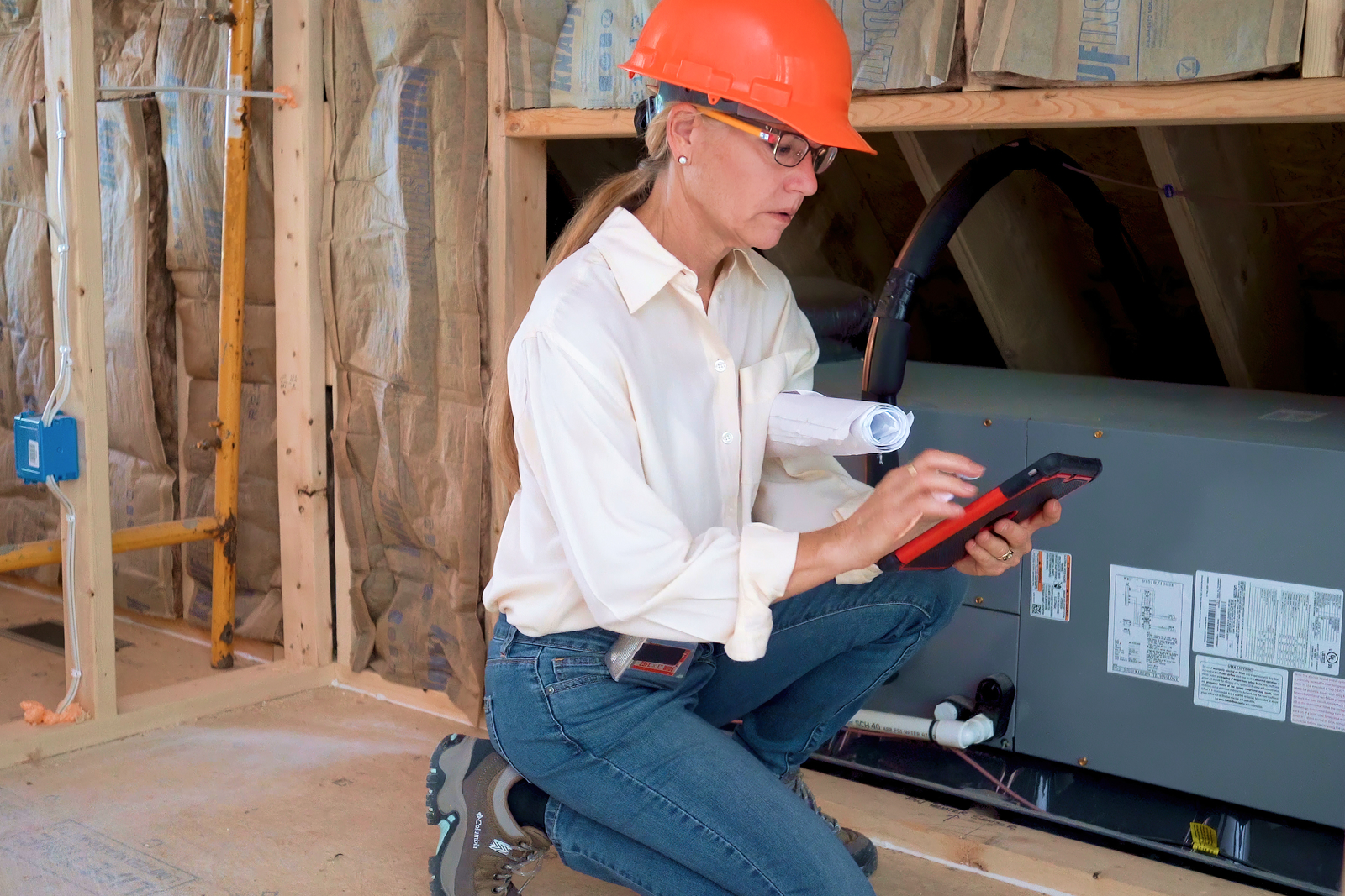 Person in white shirt and orange hard hat inspecting construction site with tablet and blueprints in hand.