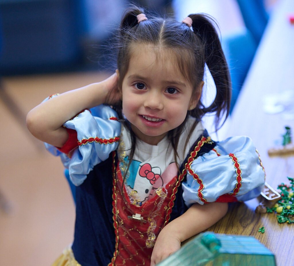 Young girl in a colorful costume with pigtails, smiling at the camera while sitting at a table indoors.