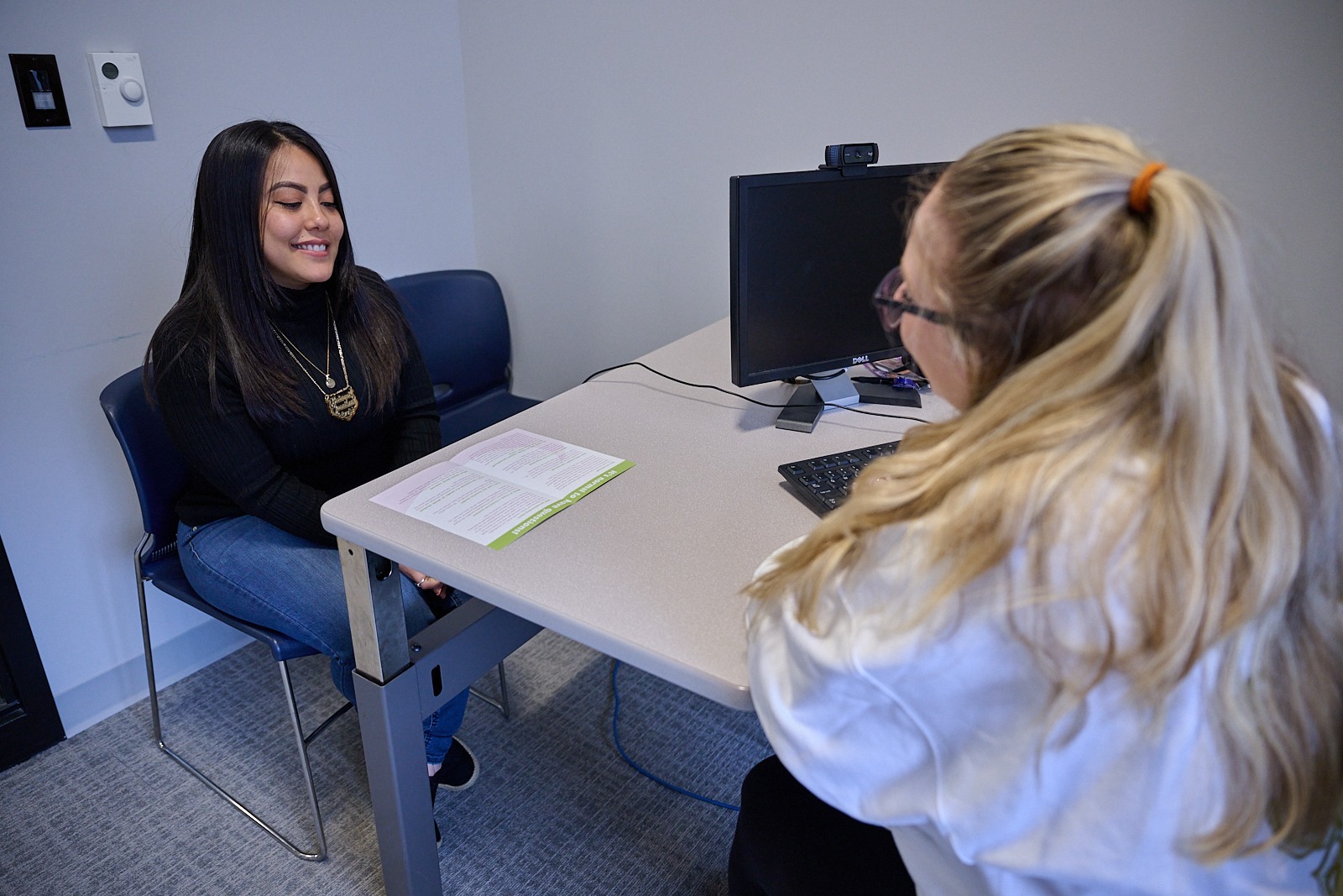Two women having a discussion in an office setting with a computer and an open book on the table.