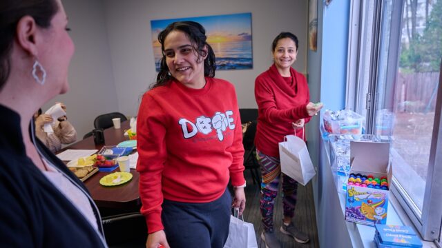 Two women in a casual office setting with one holding a gift bag and both smiling toward something out of frame.