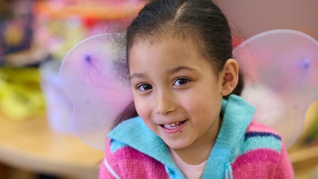 Smiling child wearing a colorful striped sweater with fairy wings in a playful, indoor setting.