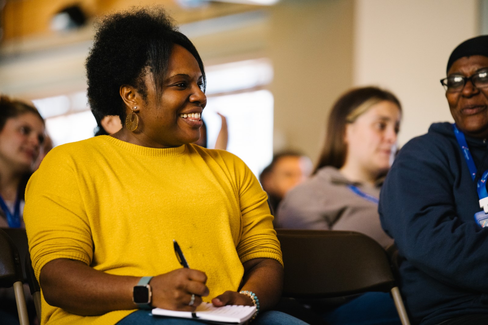 Person in a yellow shirt smiling and taking notes during a classroom session.