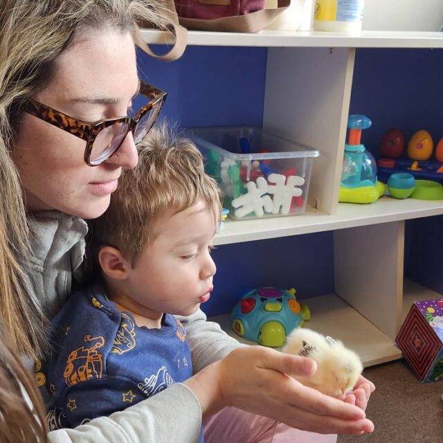 Adult and child holding a small fluffy animal, surrounded by colorful toys and shelves in a cozy setting.
