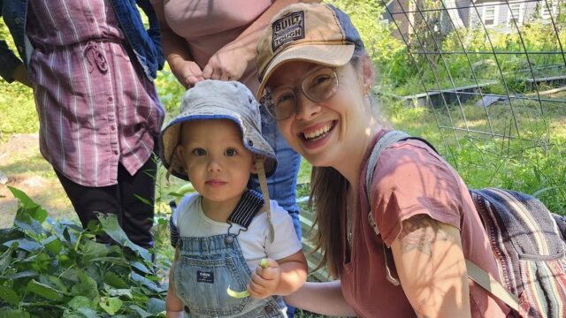 Adult and child smiling in a garden, surrounded by green plants, wearing casual clothes and sun hats.