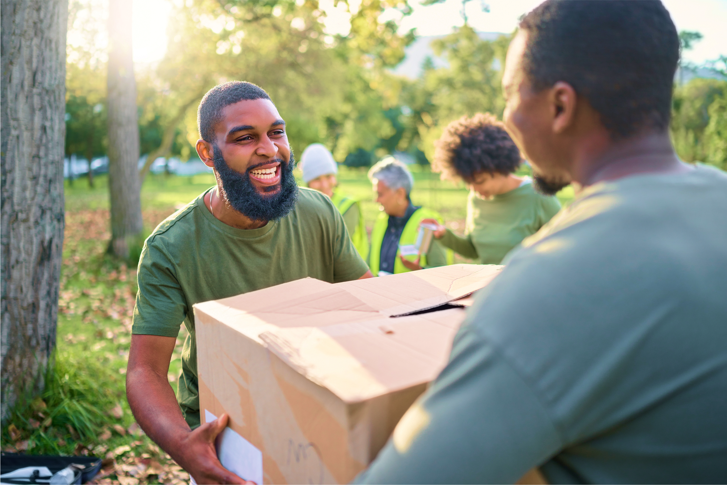 Man smiling while handing a box to another person outdoors during a community service event in a sunny park.