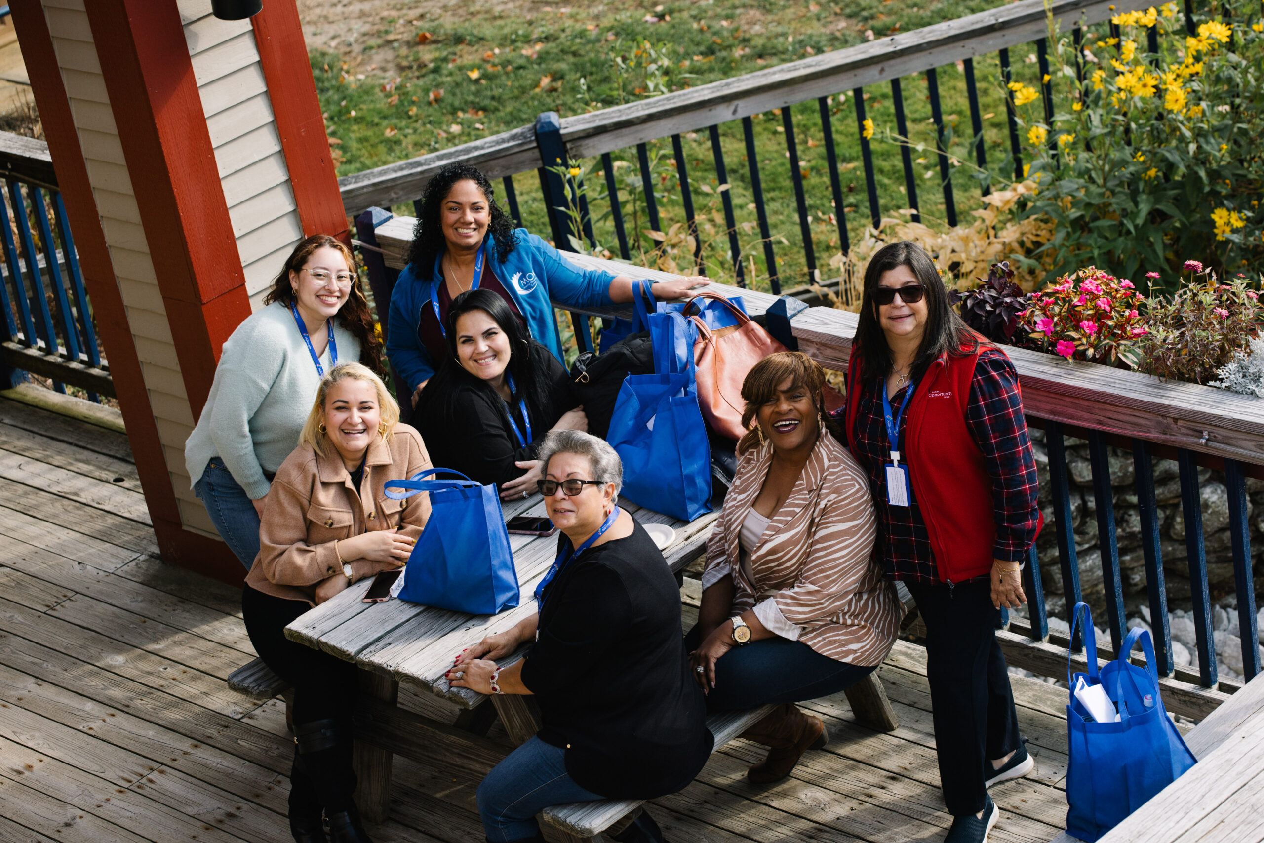 A group of people sitting around a picnic table outdoors, holding blue bags, with flowers and greenery in the background.