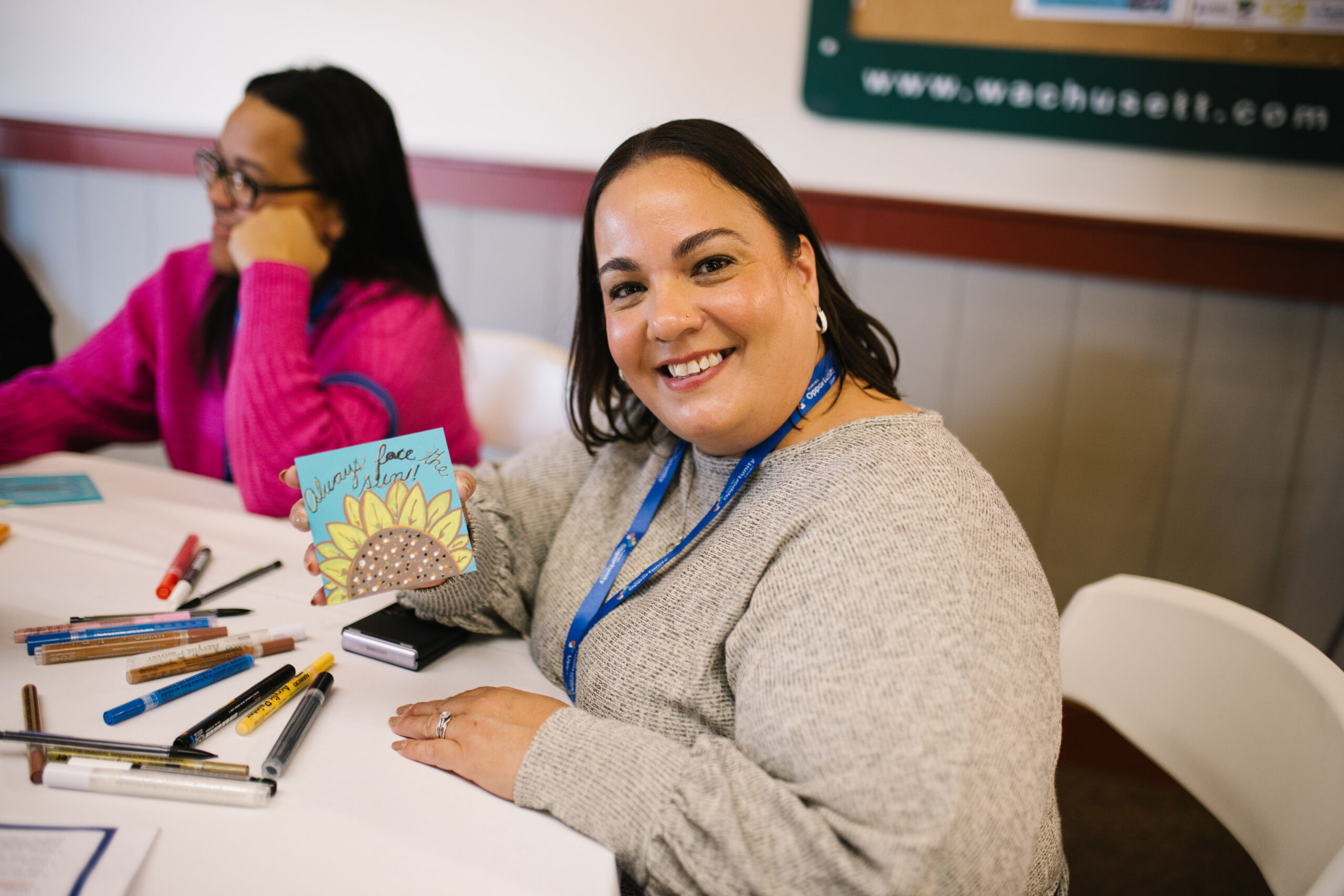 Person smiling while holding a card with a sunflower design at a table with pens and craft materials.