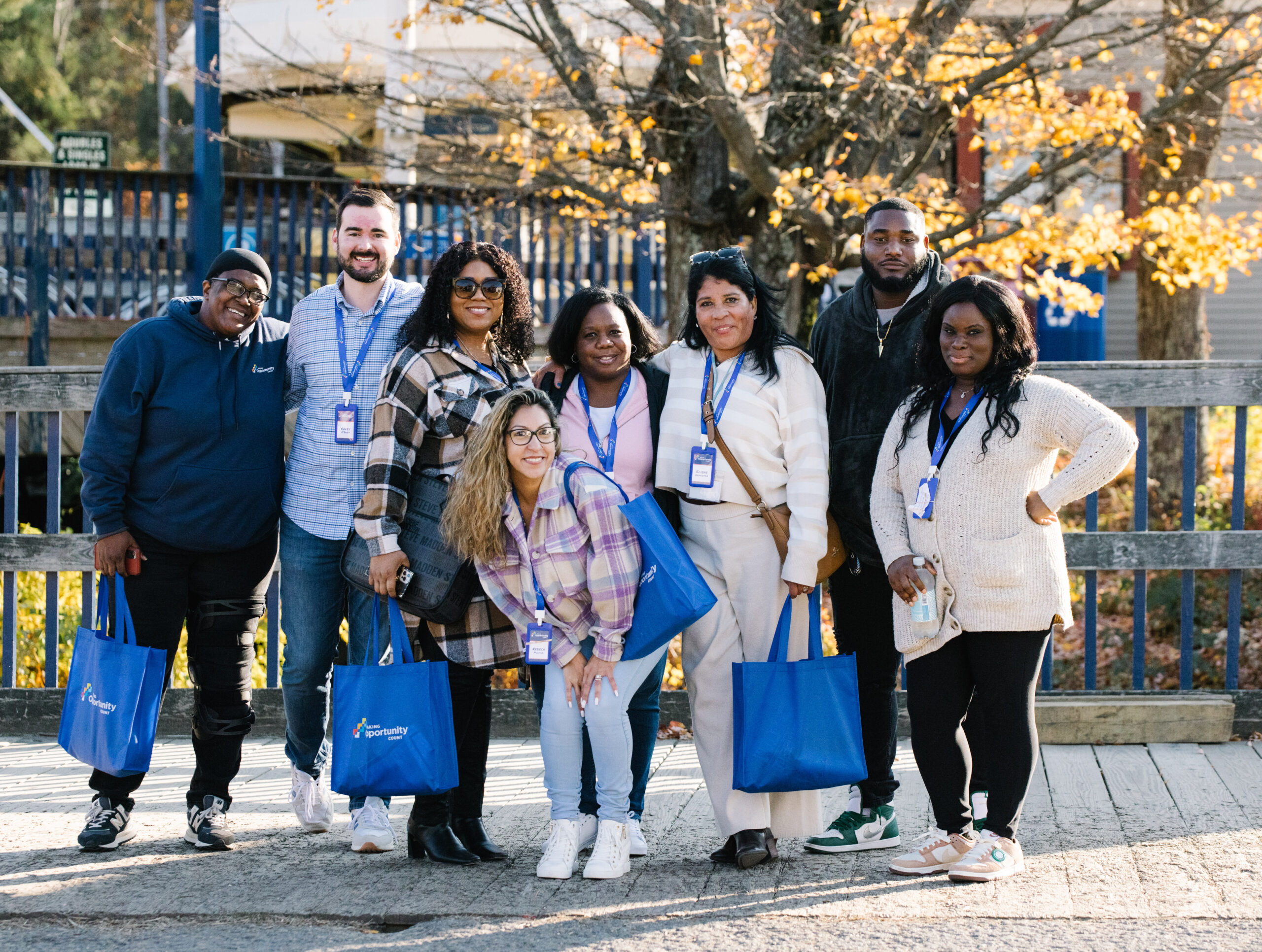 Group of people posing outside with blue bags and lanyards, standing on a path with autumn trees in the background.