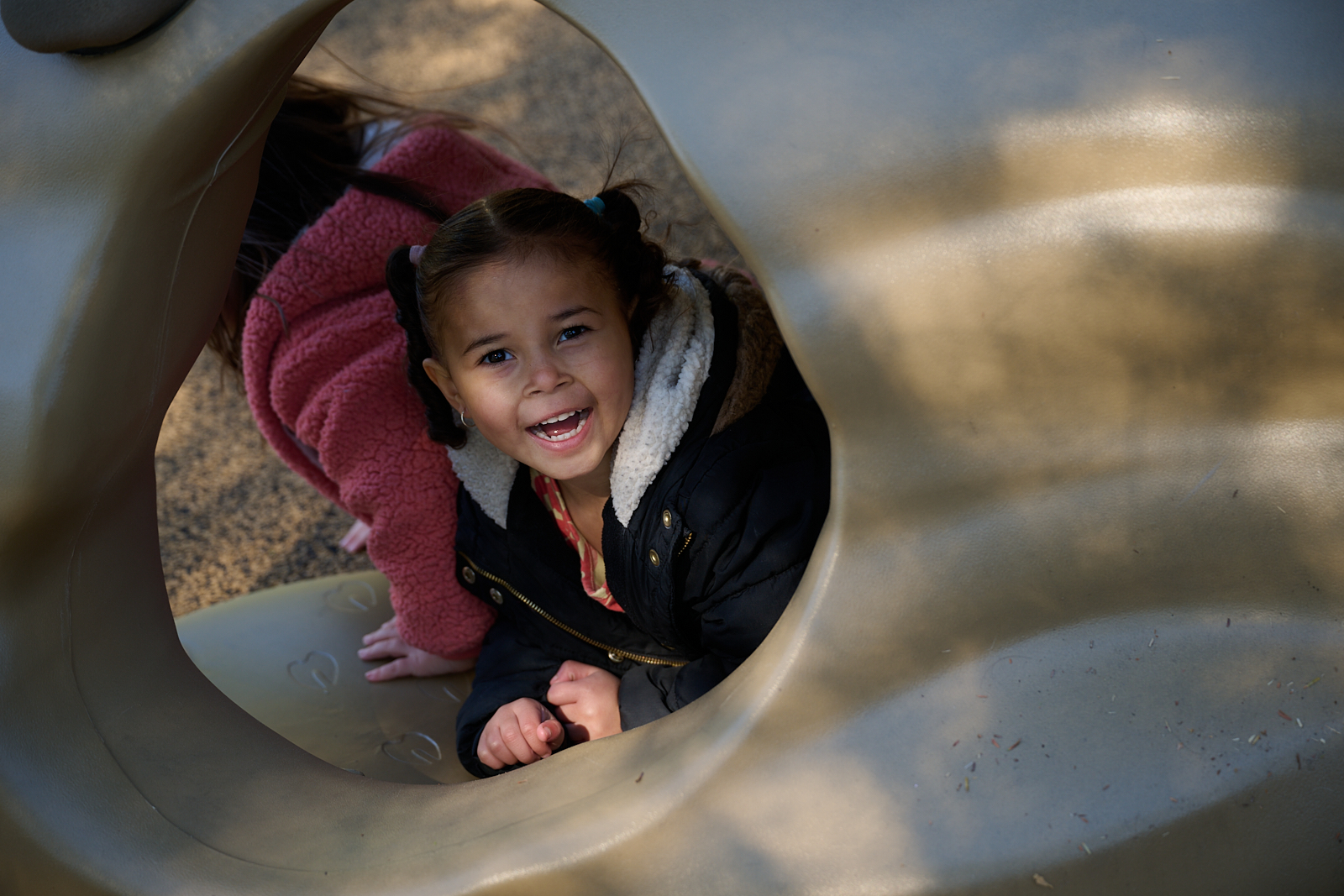 Child playing inside a playground tunnel, smiling brightly on a sunny day.