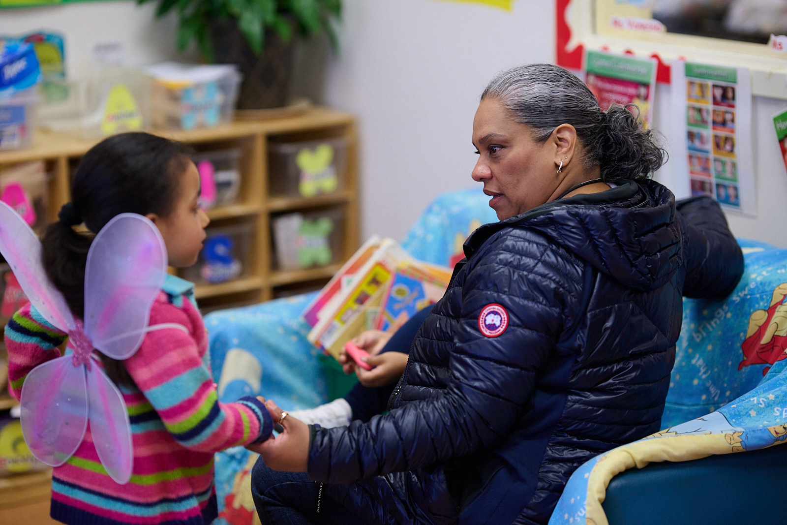 Adult holding hands with child wearing butterfly wings in a colorful classroom.