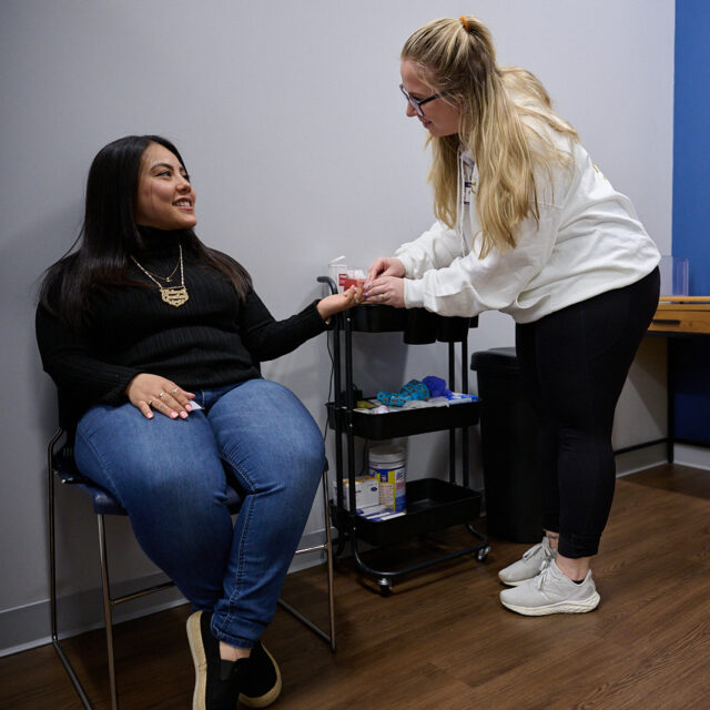 Nurse assisting a patient in a medical office with posters on the wall and a yellow examination table nearby.
