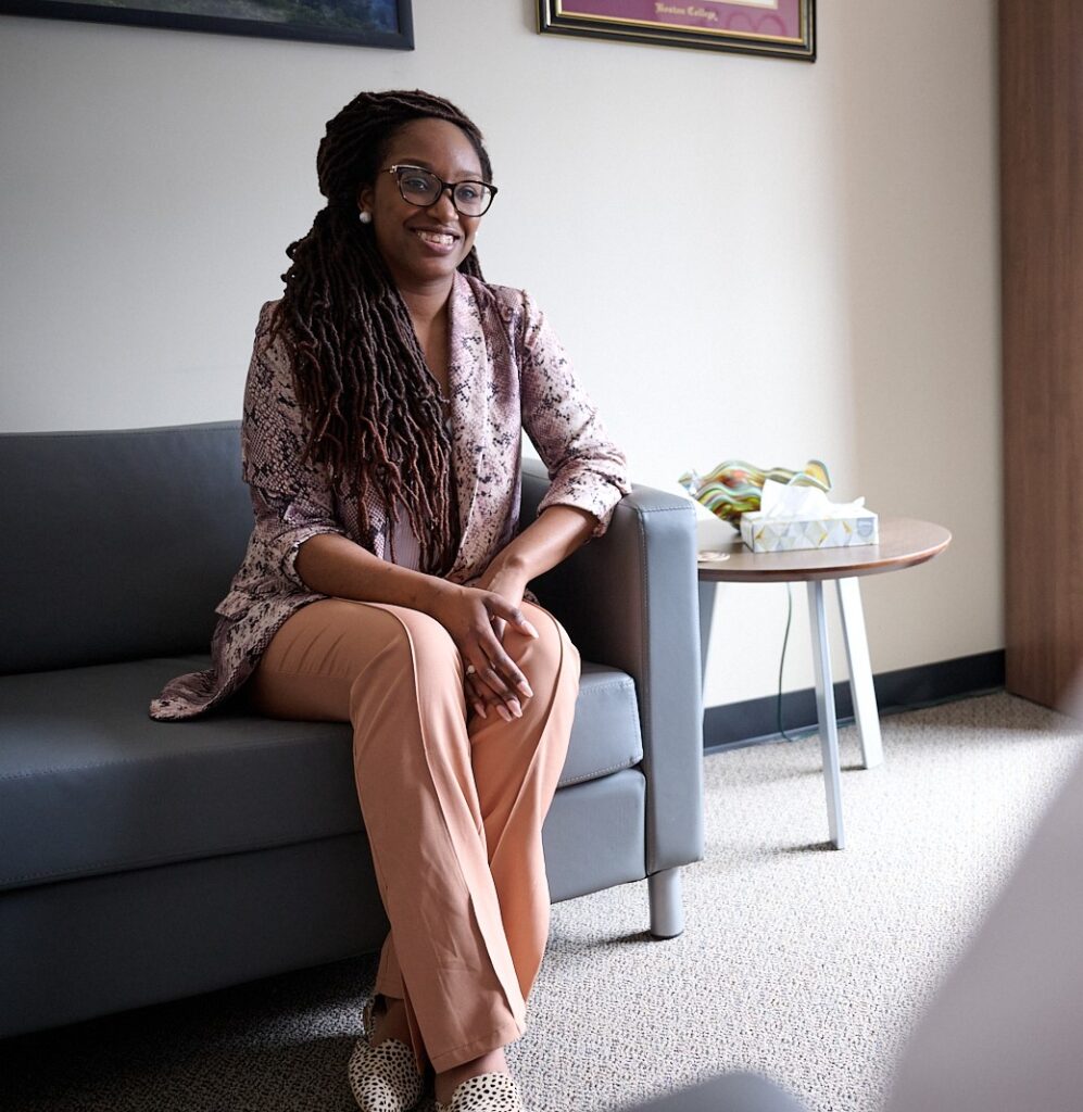 Person sitting on a couch in a well-lit room, smiling, wearing glasses and patterned clothing, with a table nearby.