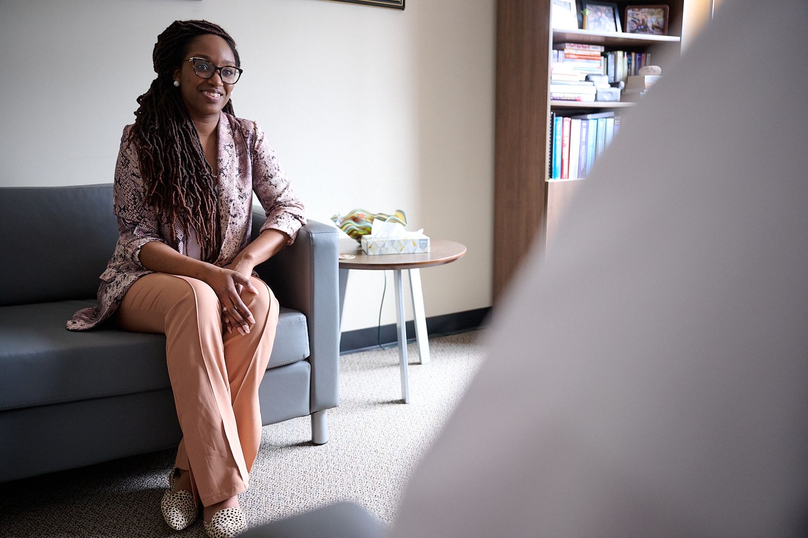 Person sitting on a gray couch in an office, engaging in conversation, with books and tissues on a nearby table.
