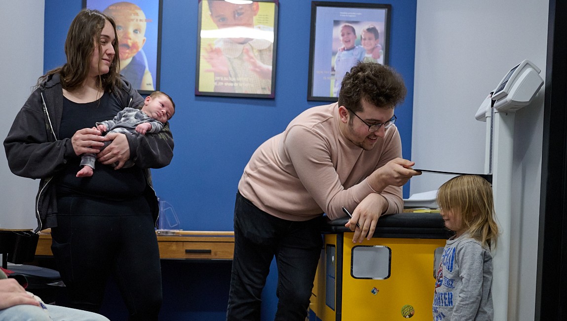 Adult measuring a child's height in a room with colorful posters, while another adult holds a baby.