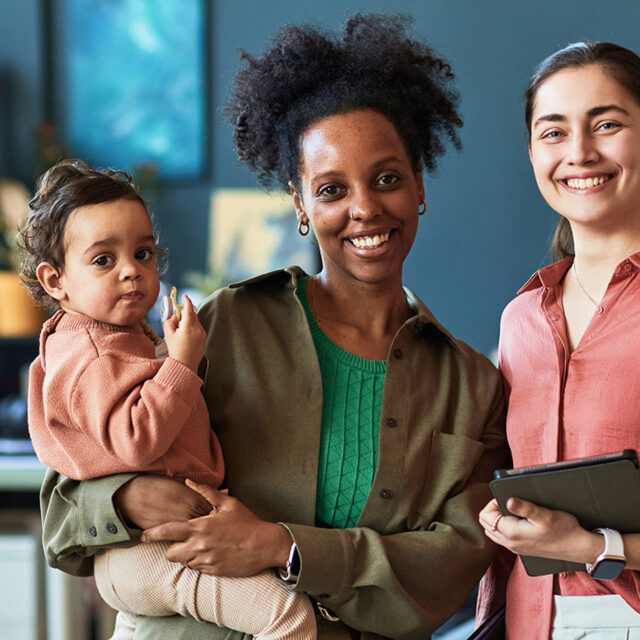 A smiling woman holds a toddler, standing next to another smiling woman in an office setting.