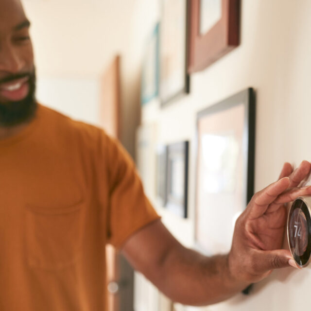 Person smiling and adjusting a digital thermostat on a wall in a warmly lit room.