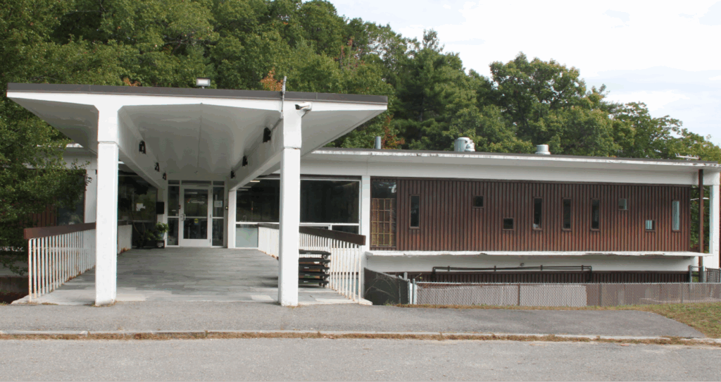 Modern building with a covered entrance, large windows, surrounded by trees in the background.