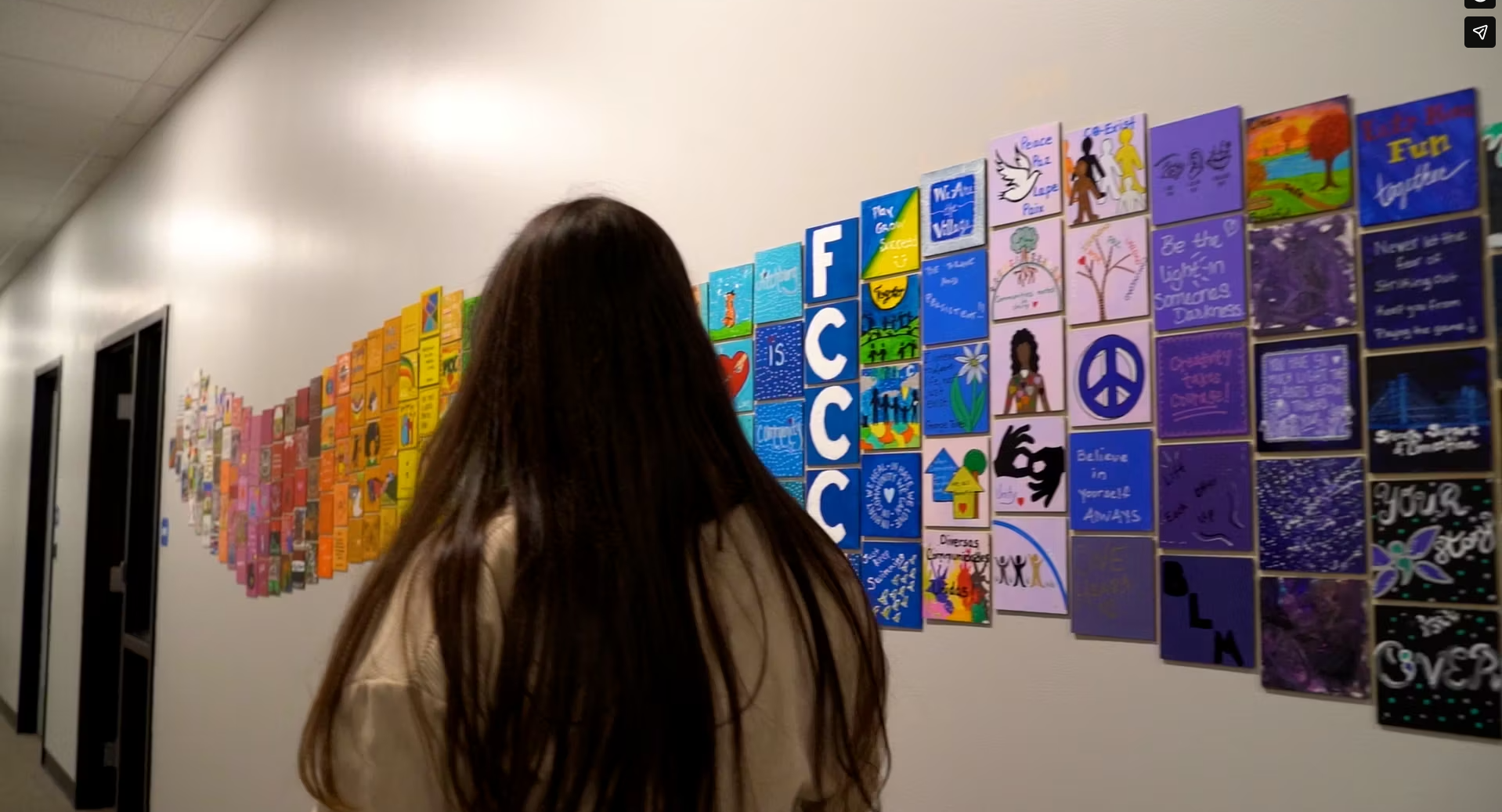 Person with long hair walking past colorful, inspiring handwritten message tiles on a wall in a hallway.