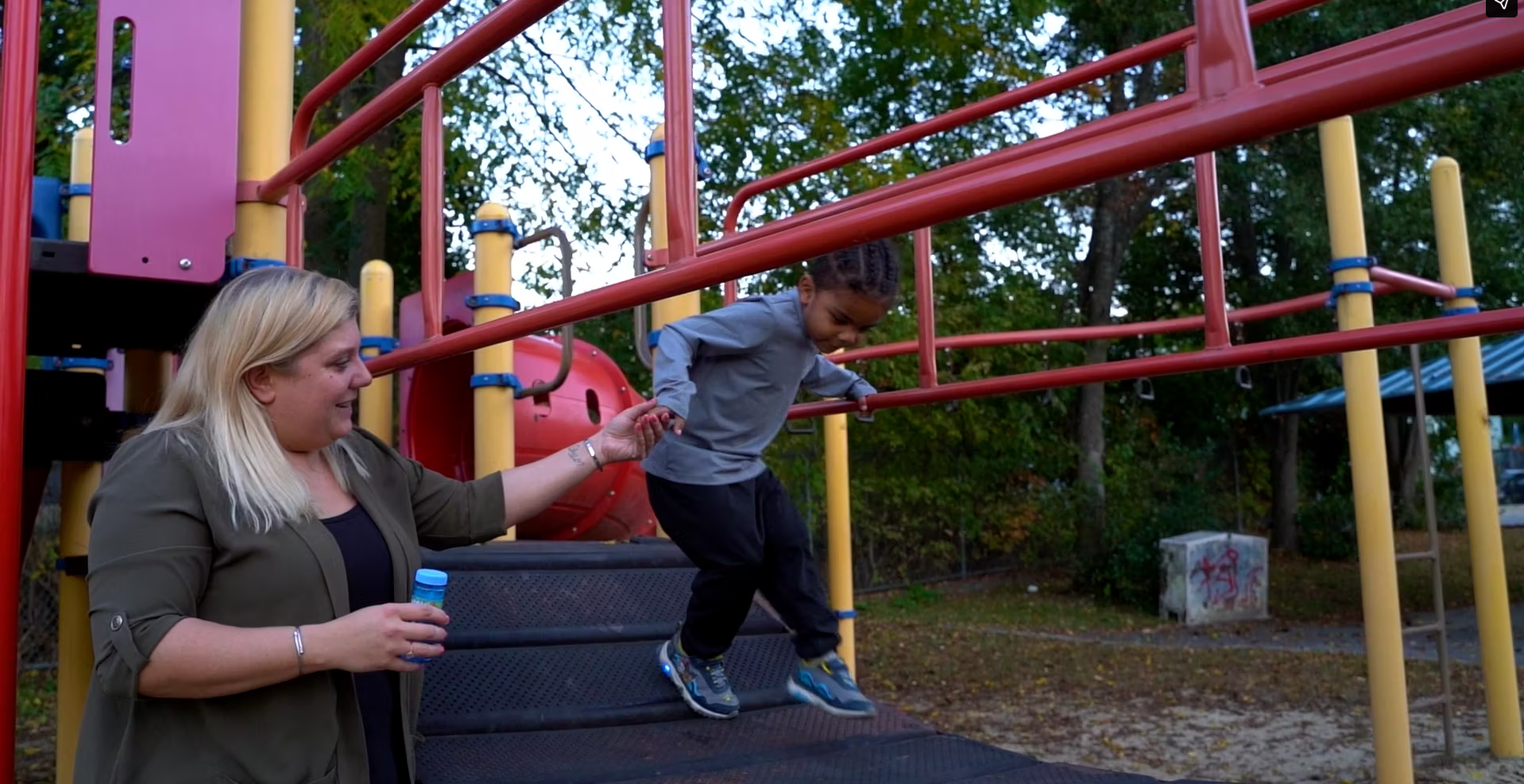 Child playing on playground slide while woman holds out hand, trees in background, sunny day in park.