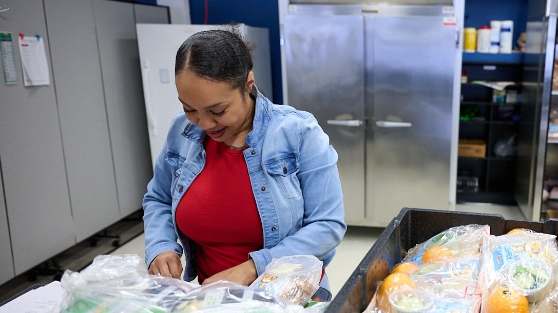 Person preparing food packages in a kitchen, surrounded by packed meals and ingredients on a table.