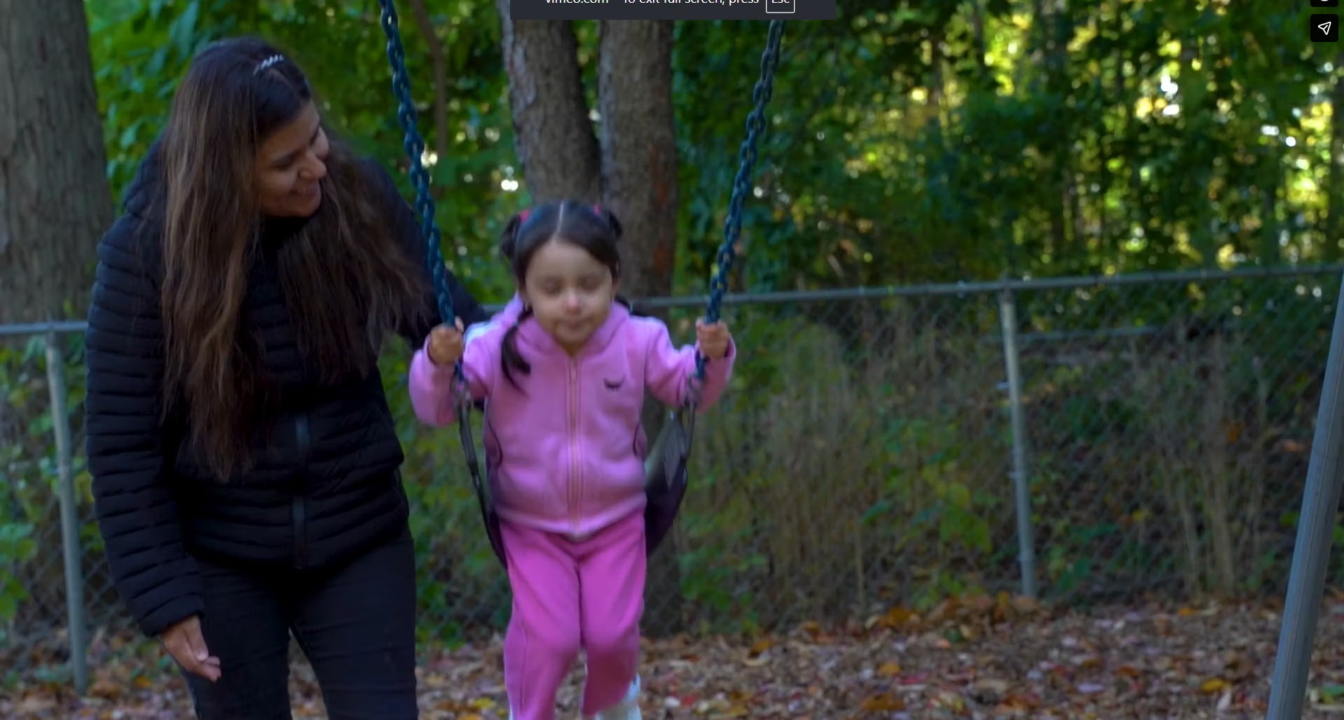 Adult guiding a child on a swing in a park surrounded by trees.