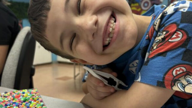 Young boy smiling brightly while leaning on a table with colorful bead art in progress.