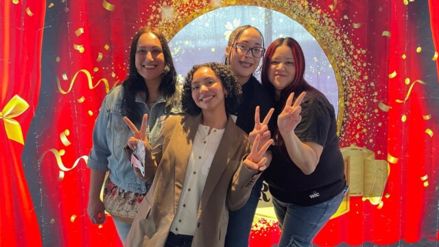 Four people smiling and posing with peace signs in front of a red and gold celebratory banner.