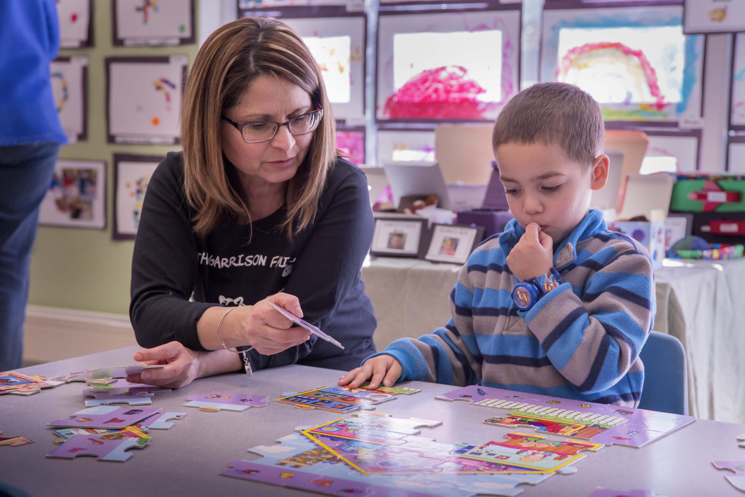 Woman helping child with a puzzle at a table in a classroom setting, surrounded by colorful artwork and crafts.