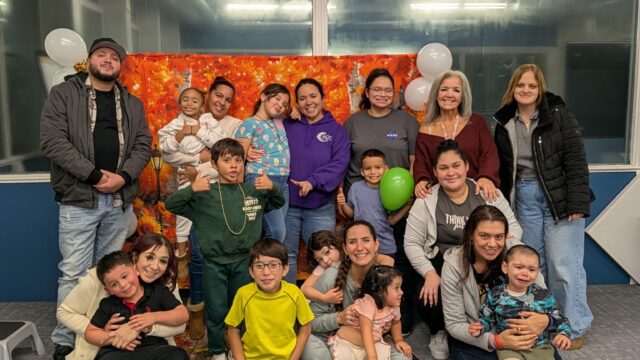 Group of children and adults smiling in front of a festive backdrop with balloons indoors.
