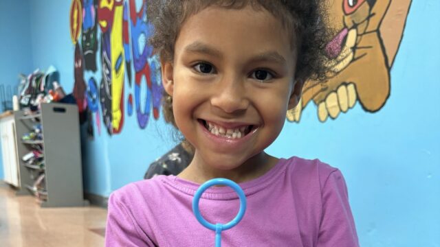Child smiling while holding a colorful pipe cleaner creation in a vibrant classroom setting.
