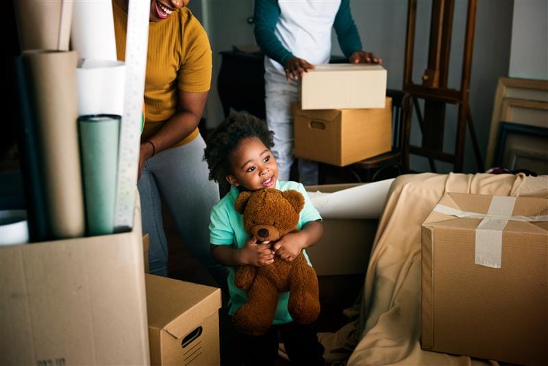 Child holding teddy bear surrounded by moving boxes indoors.