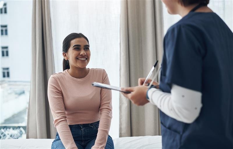 Patient sitting on examination table smiling at nurse holding a clipboard in a medical office setting.