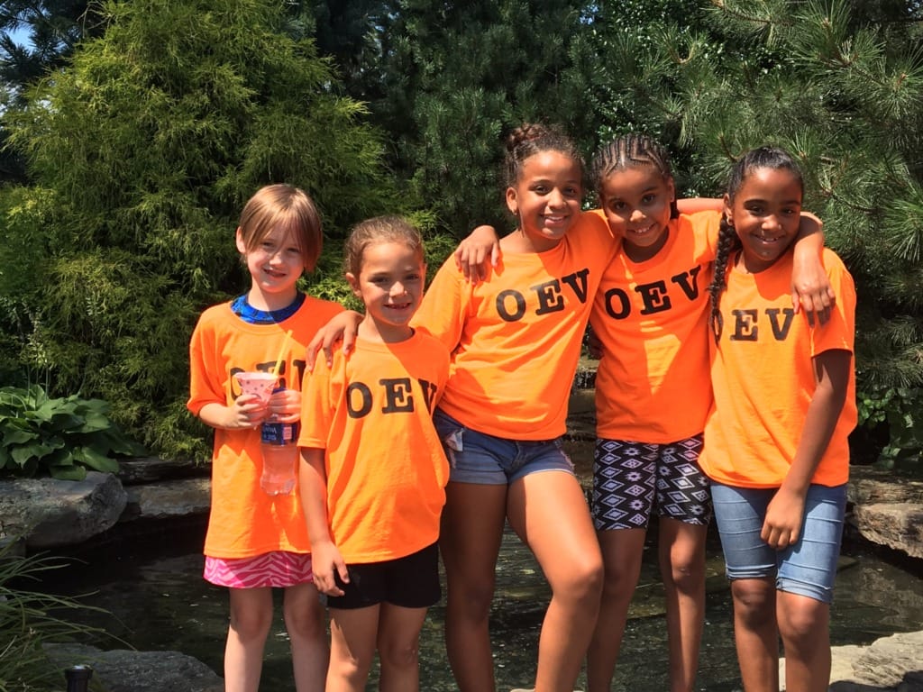 Five girls in matching orange shirts stand together smiling in front of greenery and a pond.