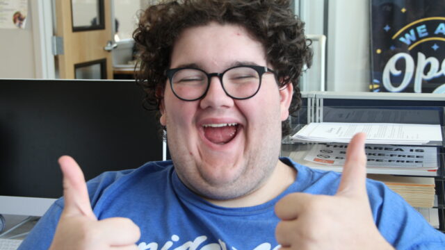 Person with glasses smiling widely and giving two thumbs up in an office setting.
