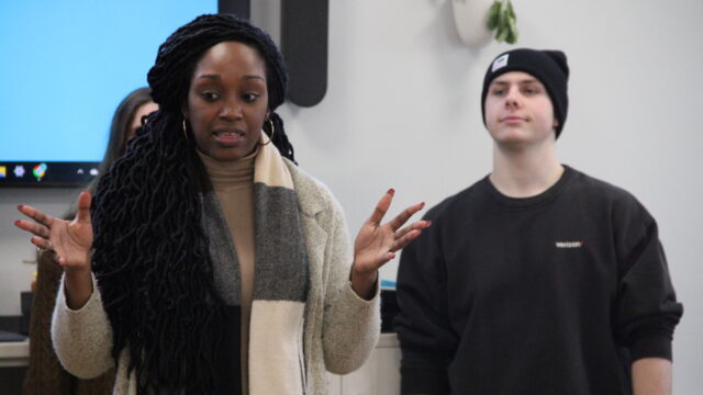 A woman speaking with expressive hand gestures while a man in a black beanie stands beside her in a room.