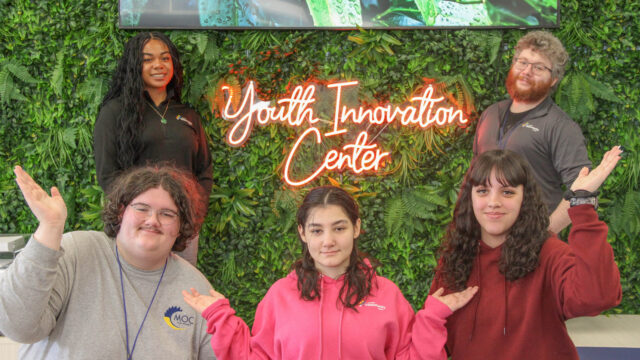 Group of five people smiling and posing in front of a "Youth Innovation Center" neon sign and greenery backdrop.