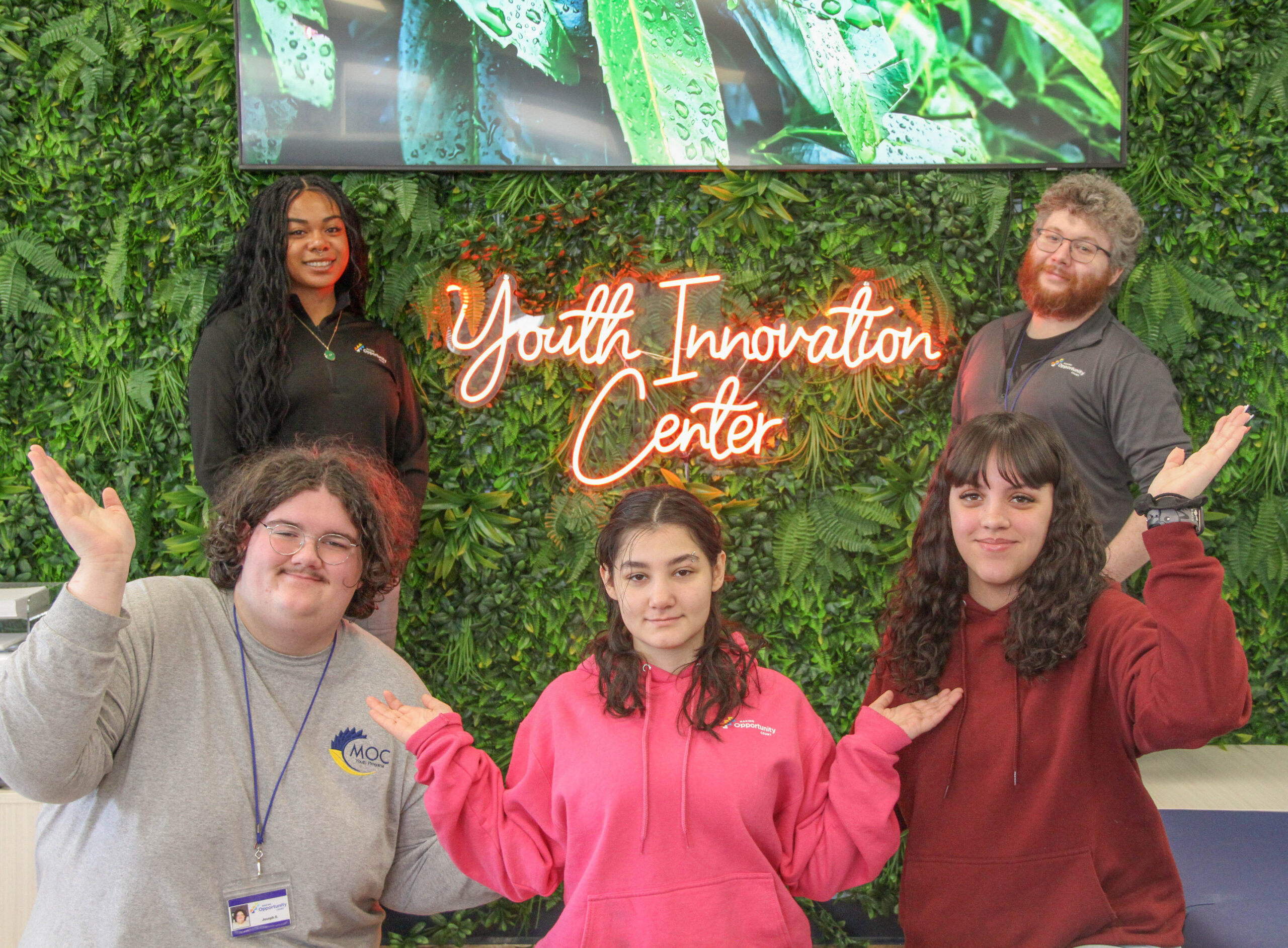 Group of five people smiling and posing in front of a "Youth Innovation Center" neon sign and greenery backdrop.