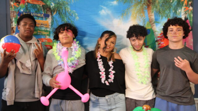 Group of friends at tropical-themed party with leis, a beach ball, and inflatable flamingo, smiling in front of scenic backdrop.