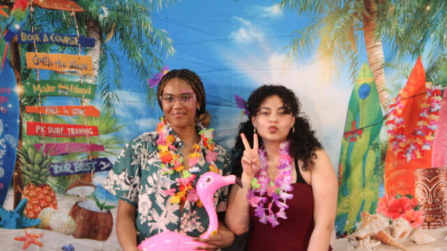 Two people with leis posing in front of a tropical-themed backdrop, one holding a pink inflatable flamingo.
